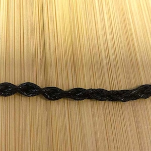 May include: Close-up of a black, braided cord. The cord has a repeating pattern of oval shapes. The background is a light brown wooden surface with vertical grain.