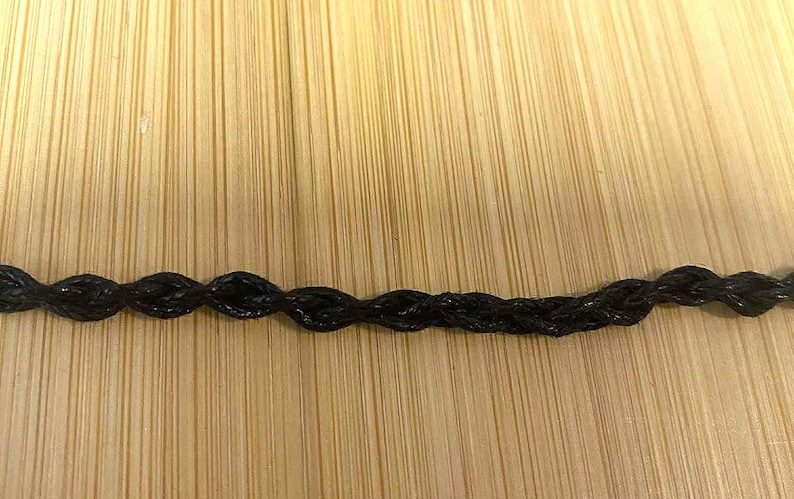 May include: Close-up of a black, braided cord. The cord has a repeating pattern of oval shapes. The background is a light brown wooden surface with vertical grain.
