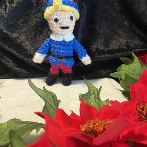 Op de afbeelding: Handgemaakte gehaakte pop van een personage met een blauw jasje, een rode broek en een blauwe hoed met gele accenten. De pop heeft een witte kraag en zwarte schoenen. De achtergrond bevat rode poinsettia's en groene bladeren.
