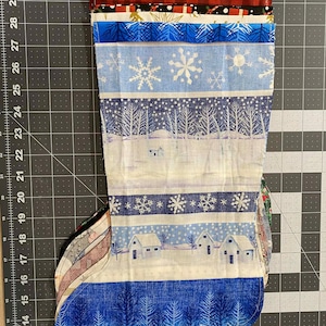 May include: A Christmas stocking with various fabric patterns. The stocking features blue and white winter scenes with snowflakes, trees, and houses. Other fabrics include red plaid and Santa Claus designs.