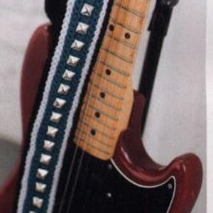 Puede incluir: Una correa de guitarra de crochet negra y blanca con tachuelas plateadas. La correa está unida a una guitarra eléctrica roja.