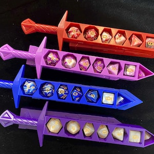 May include: Four colorful dice holders shaped like swords. Each sword is a different color: red, purple, blue, and lavender. Each sword holds a set of polyhedral dice in various colors and designs, perfect for tabletop gaming.