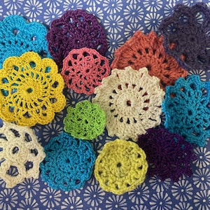 May include: A collection of colorful, handmade crochet doilies in various shades of blue, purple, yellow, green, pink, and cream. The doilies are arranged on a blue patterned surface, showcasing their intricate, circular designs.