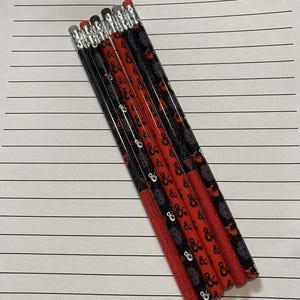 Puede incluir: Seis lápices con patrones negros y rojos, con el texto "D&D" y otros símbolos, están dispuestos en diagonal sobre papel rayado. Los lápices tienen bandas de metal plateado y gomas de borrar rosas. El papel tiene el texto "SESSION NOTES" en la parte superior.