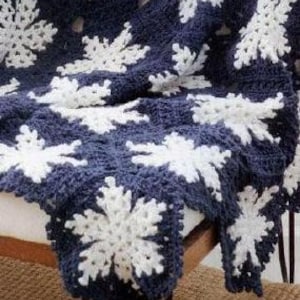 Puede incluir: Una manta de ganchillo con fondo azul marino y diseños de copos de nieve blancos.