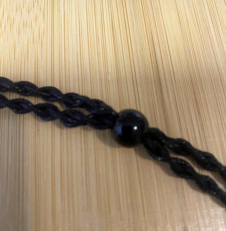 May include: Close-up of a black necklace with a glossy, round bead. The necklace is made of twisted black cord. The background is a light brown wooden surface.