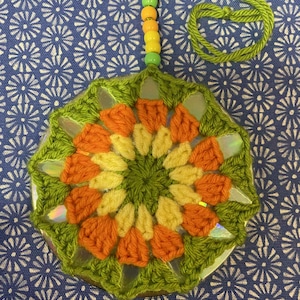 May include: A handmade, circular pendant with a green crocheted border. The center features orange and yellow crocheted petals. The pendant is attached to a green cord with orange and yellow beads. The background is a blue and white floral pattern.