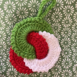 May include: A handmade crochet ornament in a spiral design. The ornament features a green outer ring, a red inner swirl, and a white section. A green loop is attached for hanging. The background is a green fabric with a white floral pattern.