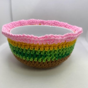 May include: A crocheted bowl cozy in brown, green, yellow, and pink stripes. The cozy is made with a crocheted stitch and fits over a white bowl.