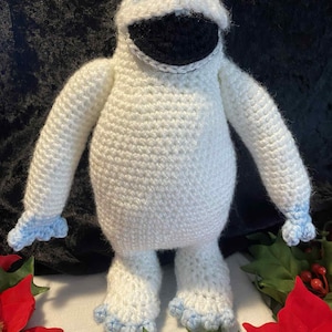May include: A handmade, white crochet abominable snowman figure with a black mouth and blue accents on the feet and hands. The snowman has a smiling expression and is posed against a dark background.