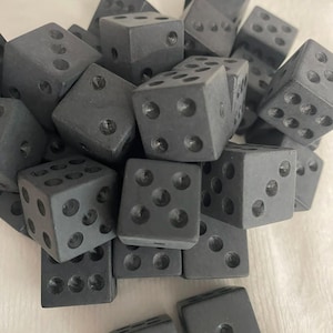 May include: A collection of dark gray dice. Each die is a cube with rounded edges and recessed circular pips. The dice are scattered, showing various numbers of pips on their faces. The overall theme is gaming and tabletop games.