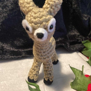 May include: A handmade crochet deer figurine in tan yarn. The deer has large, white-rimmed eyes, a white nose, and black hooves. The ears are rounded, and the body is detailed with textured stitches. The deer is standing on a white surface with green leaves and red flowers.
