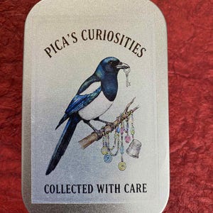 Puede incluir: Una lata de metal rectangular con una etiqueta impresa que presenta una urraca sosteniendo una llave, posada en una rama adornada con cuentas y un dedal. El texto dice "Pica's Curiosidades" y "Recogido con cuidado".