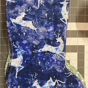 May include: A fabric Christmas stocking featuring a deep blue background with a repeating pattern of white leaping deer and small yellow stars. The stocking has a dark blue cuff at the top and is stitched along the edges with visible white thread.