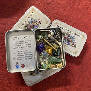 May include: A silver tin box labelled "Grizzlewick's Hoard" filled with various trinkets, including a key, dice, and a compass. The box is open, revealing its contents, and surrounded by three closed boxes with the same design. The text on the box reads "Official Trinket Box Est. 1473."