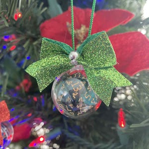 May include: A clear glass Christmas ornament with a D20 die inside, hanging from a green string. The ornament is decorated with a green glitter bow and a silver bead. The background features a Christmas tree with red poinsettias and colorful lights.