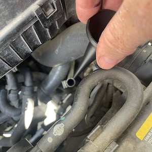 May include: A black funnel is being used to pour a liquid into a car engine. The engine components include hoses, a Nissan logo, and other mechanical parts. The image is taken from a close-up perspective.