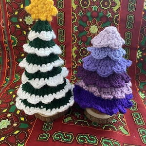 May include: Two handmade crochet Christmas trees. One is green with white trim and a yellow star topper. The other is a layered tree in shades of purple. Both trees are on small wooden bases.