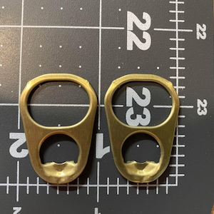 Puede incluir: Dos pestañas de abrelatas de color latón se muestran en una esterilla de corte negra con líneas de cuadrícula y números blancos. Las pestañas tienen una abertura ovalada y una abertura más pequeña y redondeada para hacer palanca.