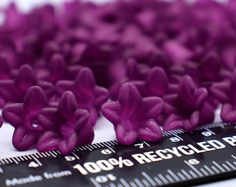 May include: A close-up of a group of purple flower-shaped plastic beads. The beads are scattered on a white surface next to a black ruler marked in centimetres. The ruler has the text "Made from 100% Recycled Plastic" printed on it.