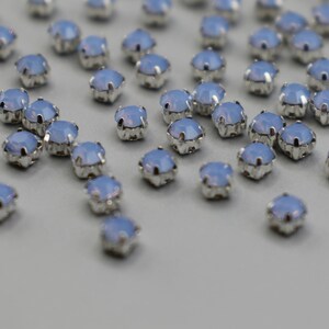May include: A collection of small, light blue gemstones with silver settings. Each gem has a faceted, star-like shape. The background is a pale grey. The gemstones are suitable for jewellery making.