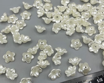 May include: A large quantity of off-white plastic flower beads. The beads are scattered on a grey surface with a ruler in the bottom right corner. The ruler is marked with numbers from 3 to 12. The ruler is made from 100% recycled plastic.