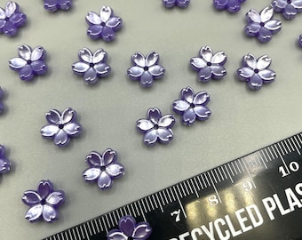 May include: A scattering of purple flower-shaped beads, some with a pearlescent finish, on a white surface. A black ruler with white markings is in the bottom right corner of the image. The ruler reads "Made from 100% Recycled Plastic".