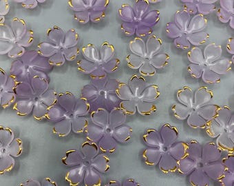 May include: A collection of purple and gold flower-shaped buttons. The buttons have a translucent, frosted appearance and are accented with a gold trim.