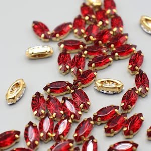 Op de afbeelding: Een verzameling rode marquise-vormige strass-steentjes met goudkleurige zettingen. Er zijn ook verschillende goudkleurige, ovale zettingen aanwezig. De strass-steentjes zijn verspreid over een lichtgrijze ondergrond, wat een sprankelend effect creëert.
