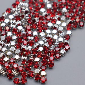 Puede incluir: Una colección de pedrería roja y plateada con un diseño facetado. Las piedras preciosas están dispersas sobre una superficie gris, creando un efecto brillante y decorativo. Estas pedrerías son adecuadas para varios proyectos de manualidades.