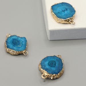 Può includere: Tre ciondoli in pietra preziosa di agata blu con bordi color oro e piccoli anelli per la creazione di gioielli. I ciondoli hanno una forma naturale e irregolare e una finitura lucida, che mette in risalto i motivi unici e il colore vibrante della pietra.