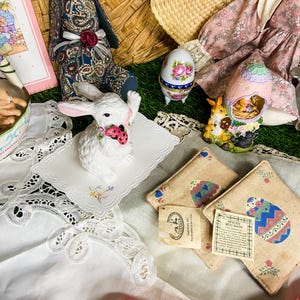 May include: A white porcelain bunny figurine holding a red ladybug, sitting on a white lace doily.  There are two brown and beige mug rugs with painted Easter eggs, a pink floral egg-shaped trinket box, and a blue and white egg-shaped trinket box.  The background includes a wicker basket, a pink floral fabric, and green grass.