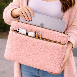 May include: A pink, fluffy laptop sleeve with a gold zip, holding a laptop, phone, charger, and pens. The sleeve has a soft, textured exterior and a zipped pocket. The laptop has the Apple logo.