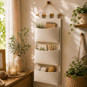 May include: A wall-mounted hanging organiser featuring three off-white canvas pockets suspended from a wooden dowel with a rope hanger. The pockets contain a spiral notebook, dried botanical stems, a rolled fabric cloth, and a wooden brush.