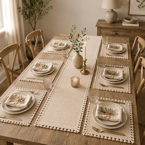 May include: A dining table set featuring a long beige runner and six matching placemats, all finished with decorative pom-pom trim. Each setting includes a white ceramic plate, a folded beige napkin tied with twine, a gold fork, and a glass.