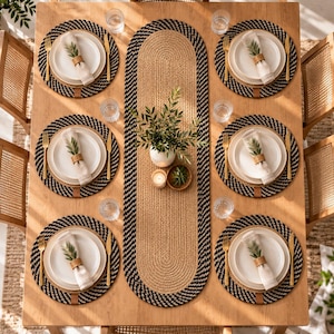 Peut inclure: Un ensemble de table comprenant six sets de table ronds tressés en fibres noires et beiges, un chemin de table ovale assorti, des assiettes en céramique blanche, des couverts dorés et des serviettes en lin avec des ronds de serviette tressés.