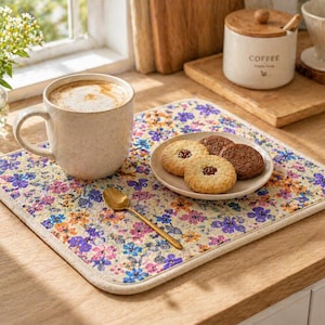 Peut inclure: Une tasse beige de cappuccino sur un set de table floral avec des biscuits. Une cuillère dorée est posée sur le tapis. Un petit vase de fleurs et un pot à café sont en arrière-plan. Le set de table a un motif floral.