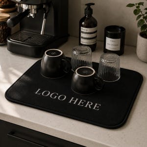 May include: A black absorbent mat with the text "LOGO HERE" in white, holding two black mugs and two clear glasses. A black espresso machine, a bottle of soap, and a black candle are in the background.