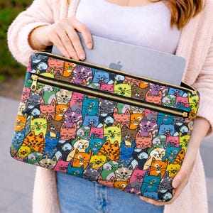 May include: A laptop sleeve with a vibrant pattern of cartoon cats in multiple colours. It has a gold zip closure. The sleeve is held by a person, and contains a laptop with the Apple logo.