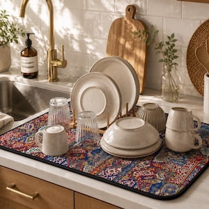 Peut inclure: Une scène de cuisine avec un tapis de séchage de vaisselle à motifs accueillant des assiettes, des bols, des verres et des tasses beiges et blancs. Le tapis présente un motif rouge, bleu et or. Un robinet doré et une petite plante en pot sont également visibles.