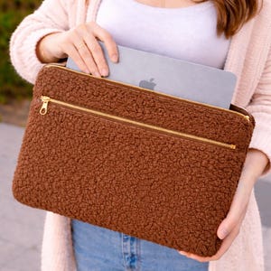 May include: A brown laptop sleeve with a textured, fuzzy exterior. The sleeve has a gold zipper closure and a gold zipper pocket on the front. A silver laptop with an Apple logo is partially visible inside the sleeve.