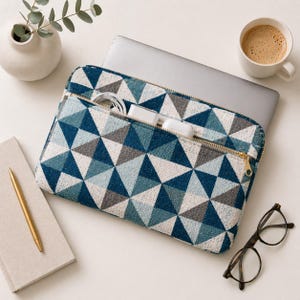 May include: A blue, gray, and white geometric patterned laptop sleeve with a gold zipper. The sleeve is open, revealing a laptop and charging accessories. A gold pen, notebook, glasses, and a cup of coffee are also on the white surface.