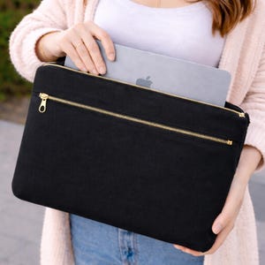 May include: A black rectangular laptop sleeve featuring a gold-toned metal zipper closure across the top and a secondary horizontal zippered pocket on the front. A silver laptop with an Apple logo is partially inserted into the main compartment.