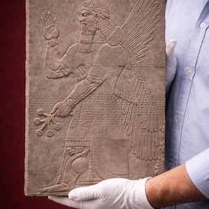 May include: A carved stone relief depicting a winged figure with a detailed beard, headdress, and intricate clothing. The figure holds a floral arrangement in one hand and raises the other. The relief is held by gloved hands.