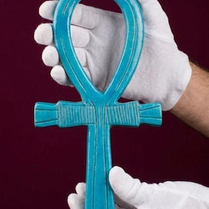 Può includere: Un simbolo ankh color turchese brillante con una finitura liscia e lucida, caratterizzato da dettagli a linee verticali incise lungo la traversa e lo stelo centrale. L'oggetto ha la forma di una croce con una parte superiore ad anello.