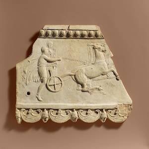 May include: A light-colored, rectangular relief sculpture depicting a chariot race. The relief shows a figure driving a chariot pulled by a horse. The top and bottom edges feature decorative molding. The sculpture is chipped along the edges.