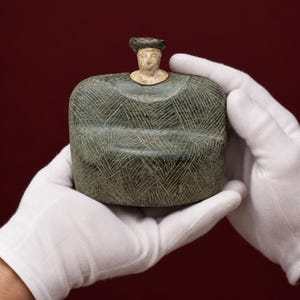 Può includere: Una scultura in pietra verde scuro di una figura seduta con una testa liscia di colore chiaro e un corpo piatto e largo. Il corpo presenta un intricato motivo a tratteggio incrociato inciso che copre l'intera superficie, creando texture.