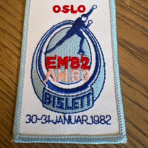 May include: A rectangular embroidered patch with a white background and light blue border. It features a dark blue ice skater silhouette, the red text OSLO, EM'82, AW'83 in light pink, the blue text BISLETT, and the date 30-31 JANUAR 1982 in blue.