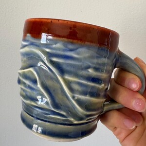 May include: Handmade ceramic mug with a unique textured design. The mug features a dark red rim, transitioning to blue and cream tones on the body. The handle is a matching blue-grey. A functional and decorative piece for enjoying beverages.