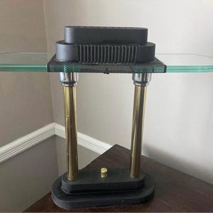 May include: A vintage table lamp with a clear glass shelf and a black top. The lamp has two gold-colored metal supports and a black base. The lamp is on a dark brown wooden surface.